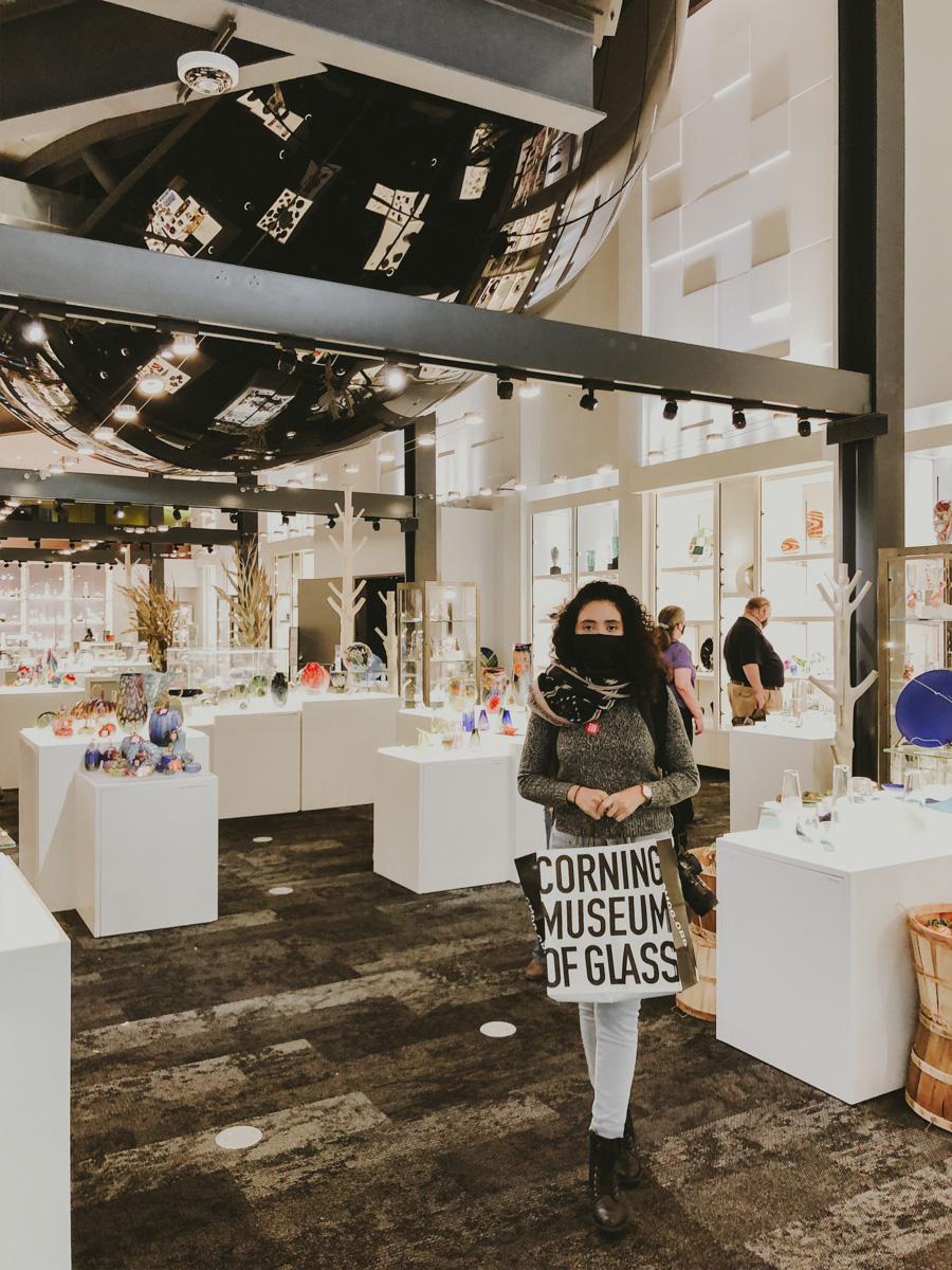 A woman inside the Corning Museum of Glass surrounded by glass items and holding a shopping bag
