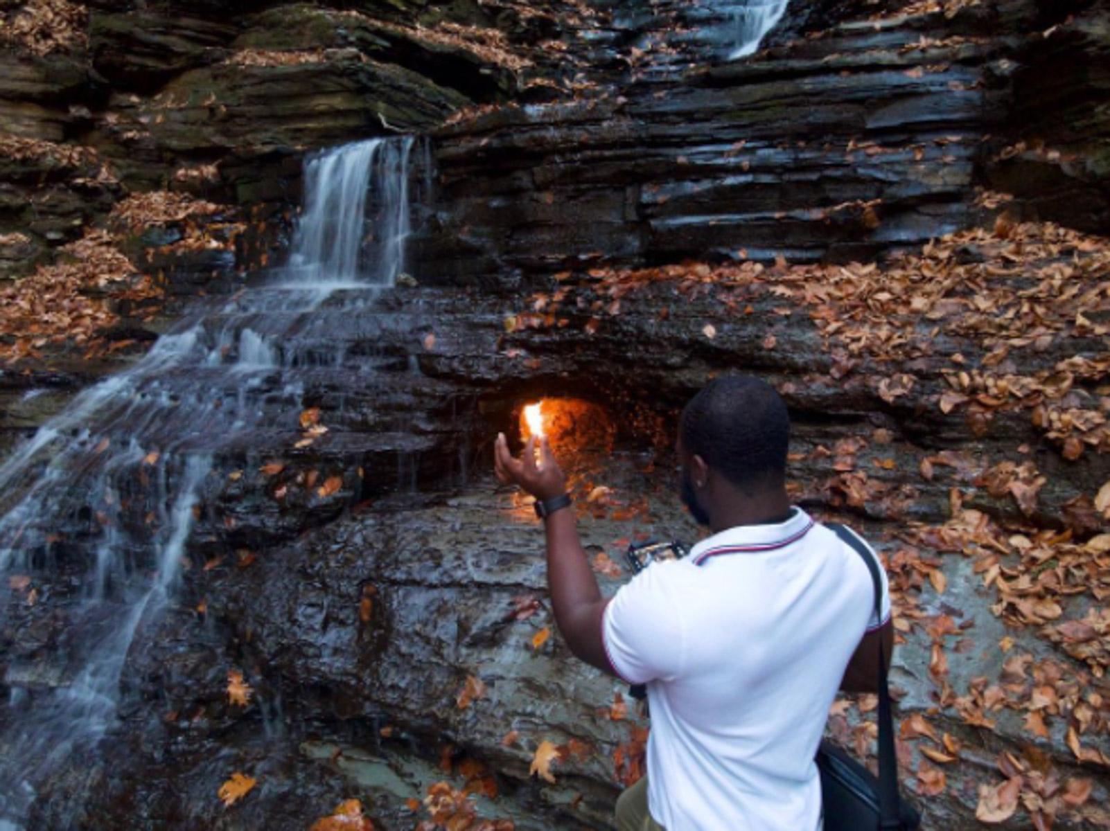 A man facing backwards looking at a waterfall and a small grotto with a flame in it.