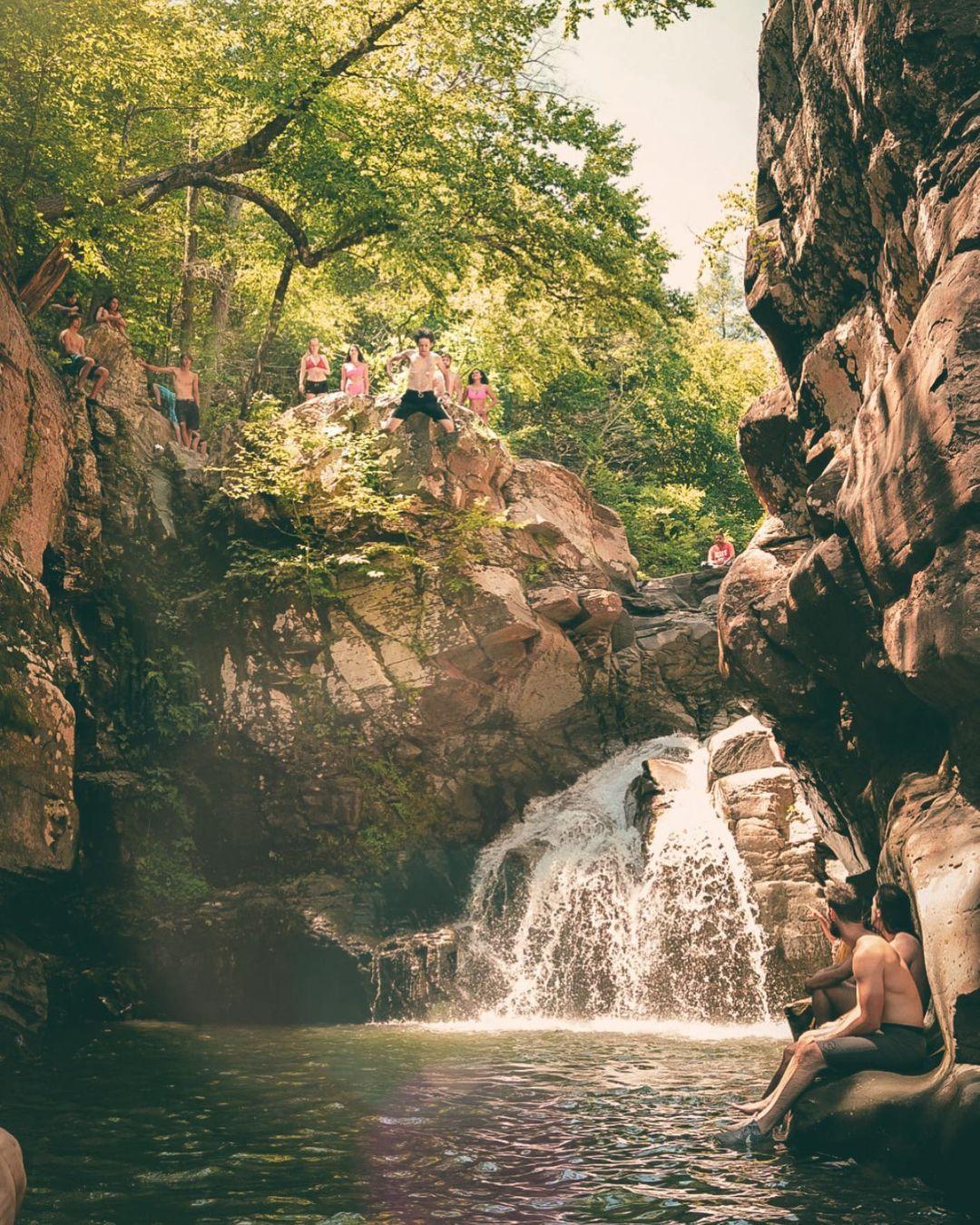 People standing and jumping off a gorge to a natural swimming pool in front of a waterfall.