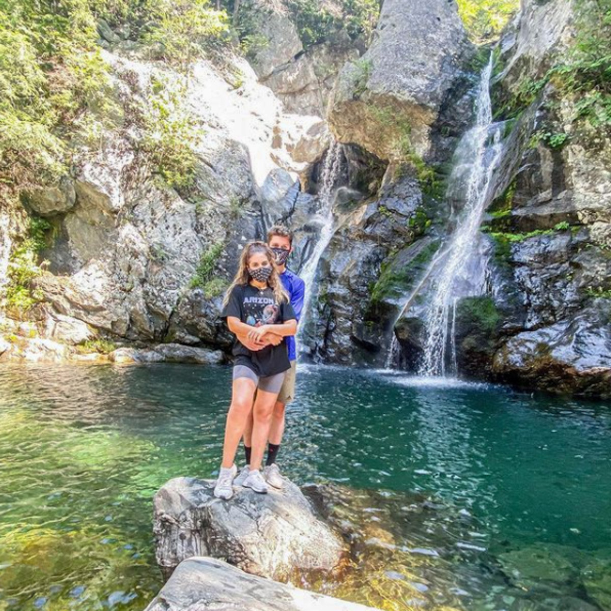 A man and a woman standing on a rock in front of a waterfall.