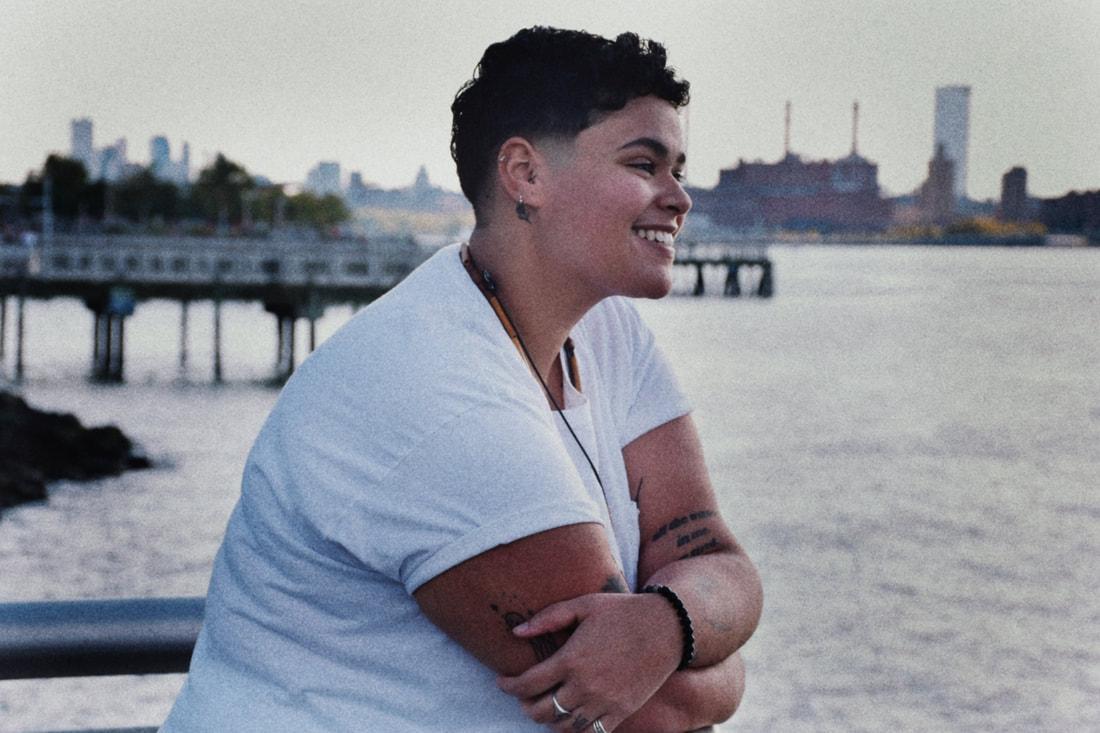 A woman with short hair and wearing a white shirt standing outdoors in front of a body of water in a city.