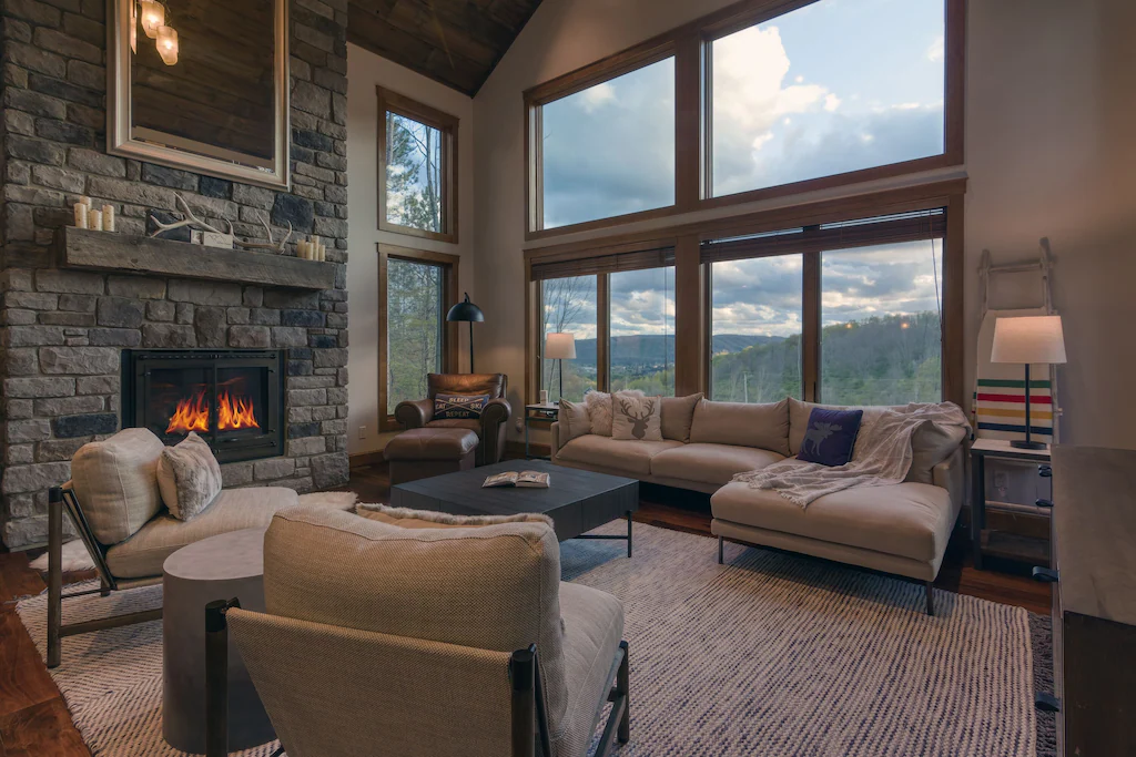 The living room area of a luxurious cabin with furniture, fireplace, and windows with mountain views.