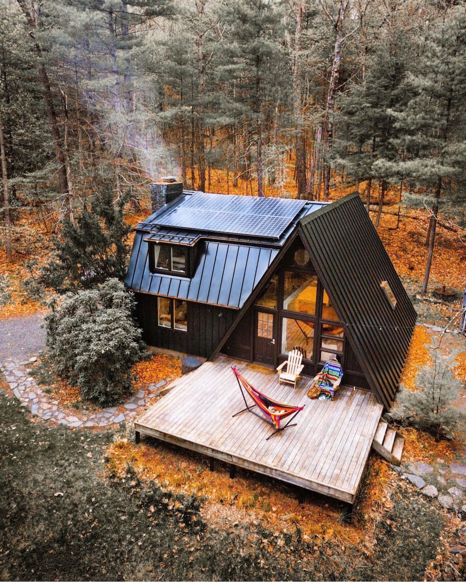 A black cabin with chimney and with hammock and chairs outside surrounded by trees.