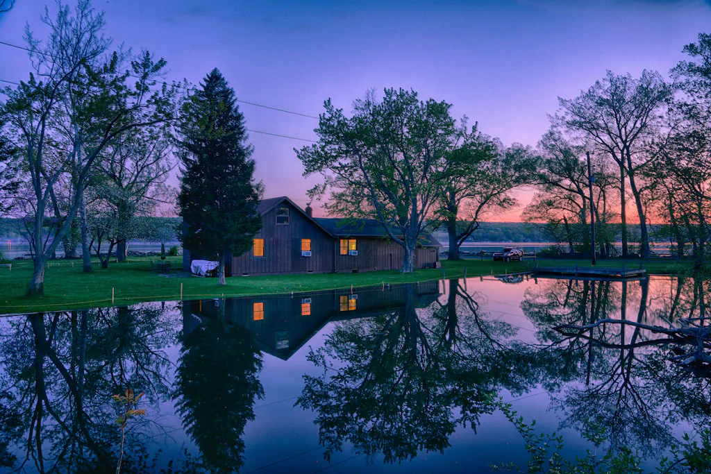 A cabin near Cayuga Lake with trees reflecting on a body of water and purple sky.