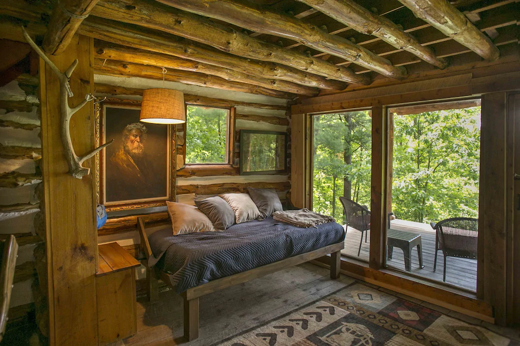The inside of a cabin with carpet, chair, pillows, and painting and with a door that leads to an outdoor sitting area.