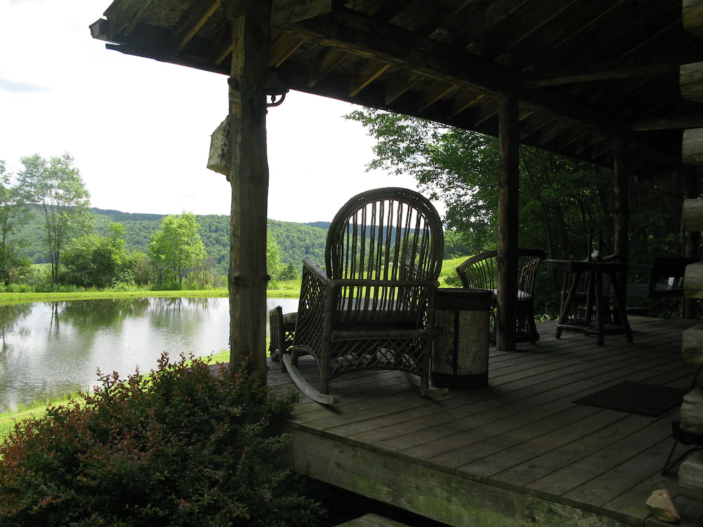 Wooden chairs at the outdoor area of a log cabin with a view of a pond.