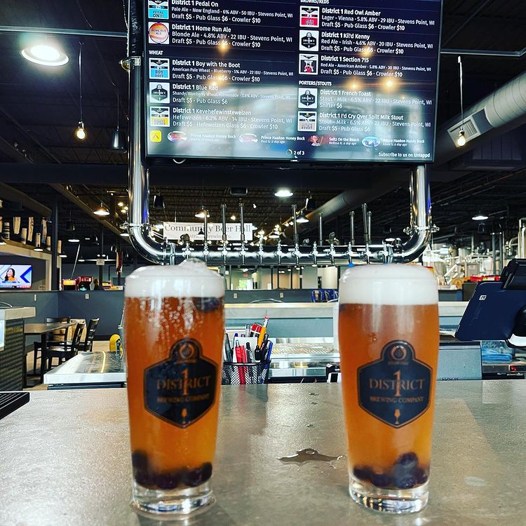 Two full glasses of beer with brewery's logo and brewery's interior in the background.