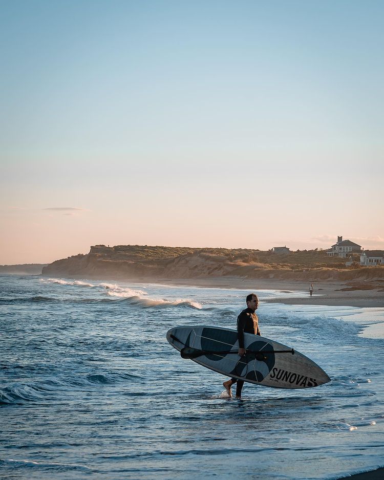 Man carrying surfing board and getting out of the water. In the distance beautiful house and sandy Beach. 