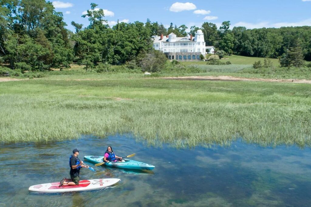 Man and a woman kayakoing next to each other. In the background a lake house and beautiful trees and meadow.
