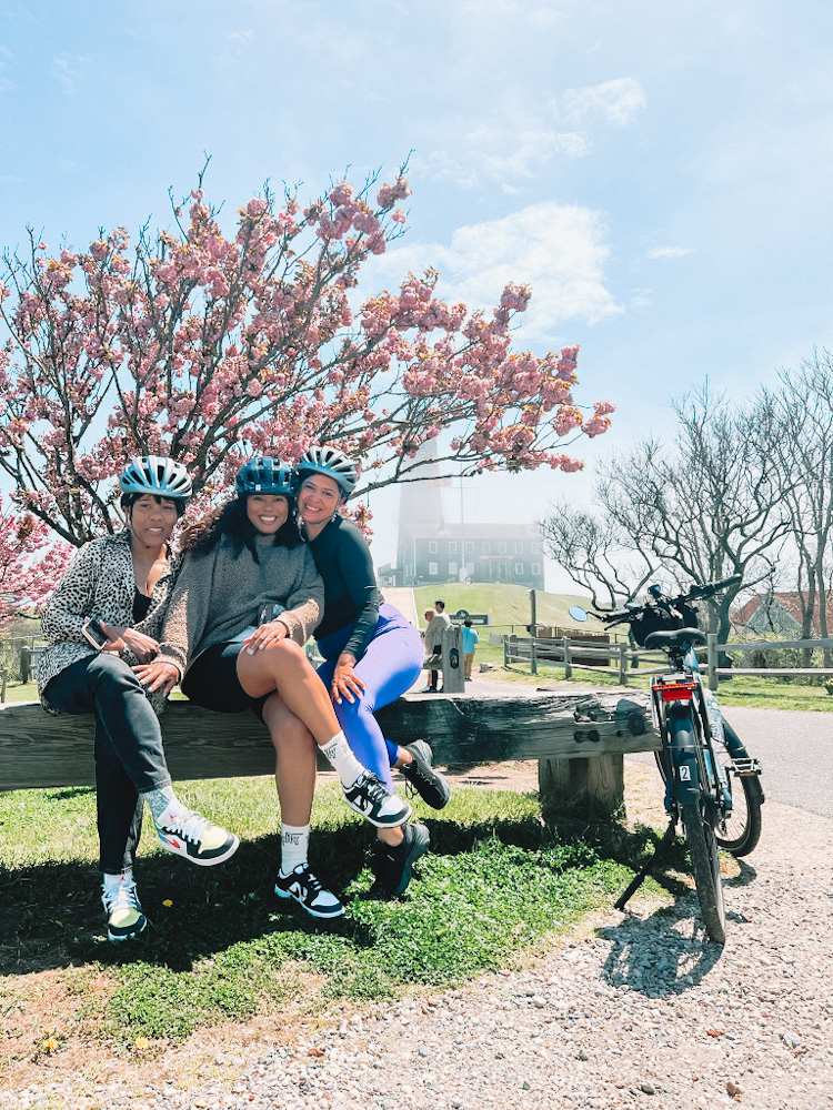 Three women sitting on a bench in front of a tree and a bike leaning on the same bench.