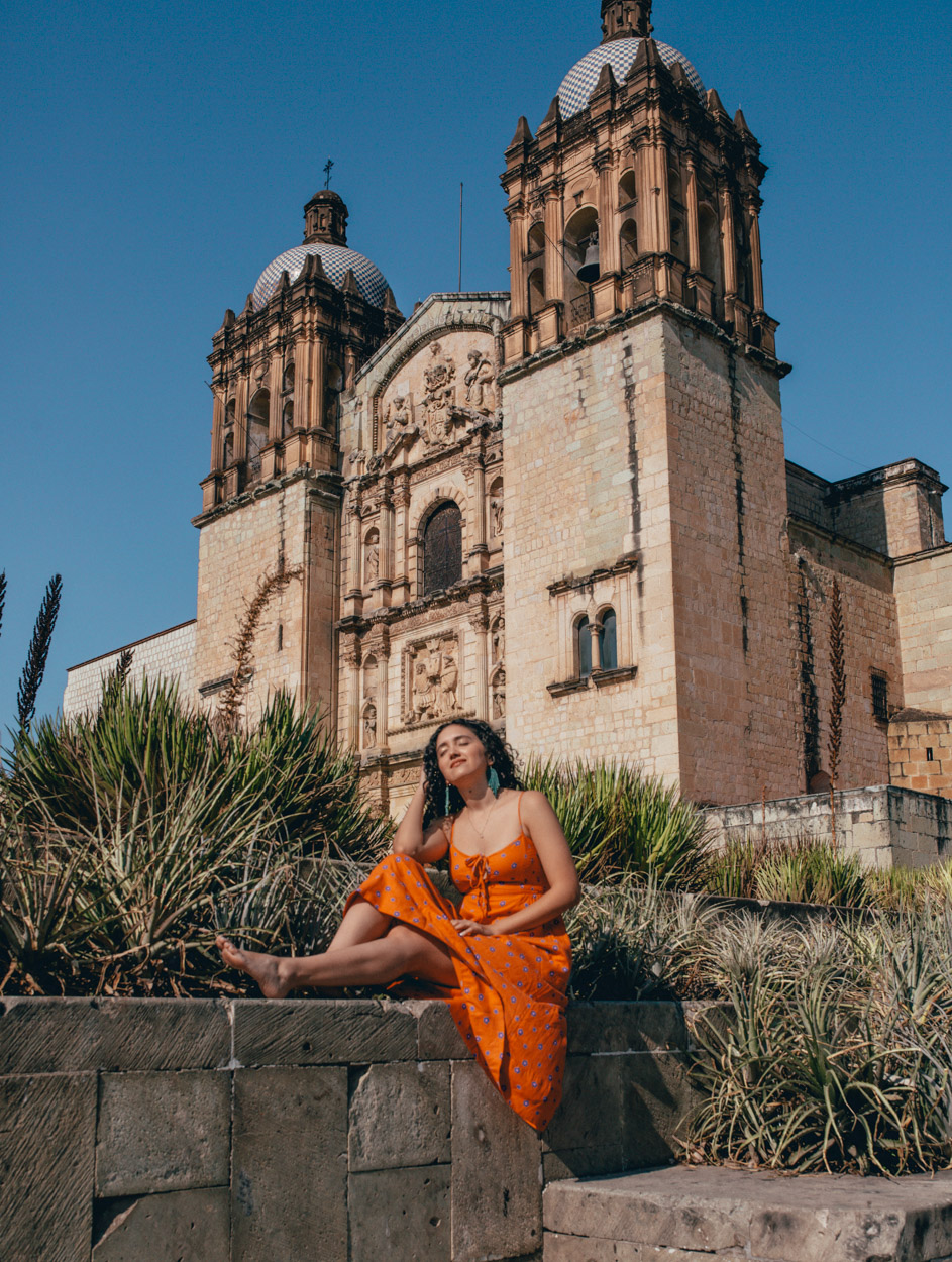 A yellow woman in orange dress sitting in front of a church in Oaxaca.