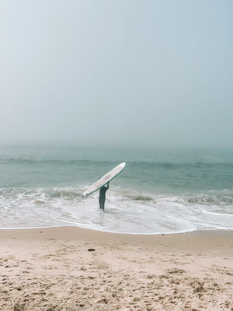 A person holding a surfing board above them at the beach in Montauk day trip.