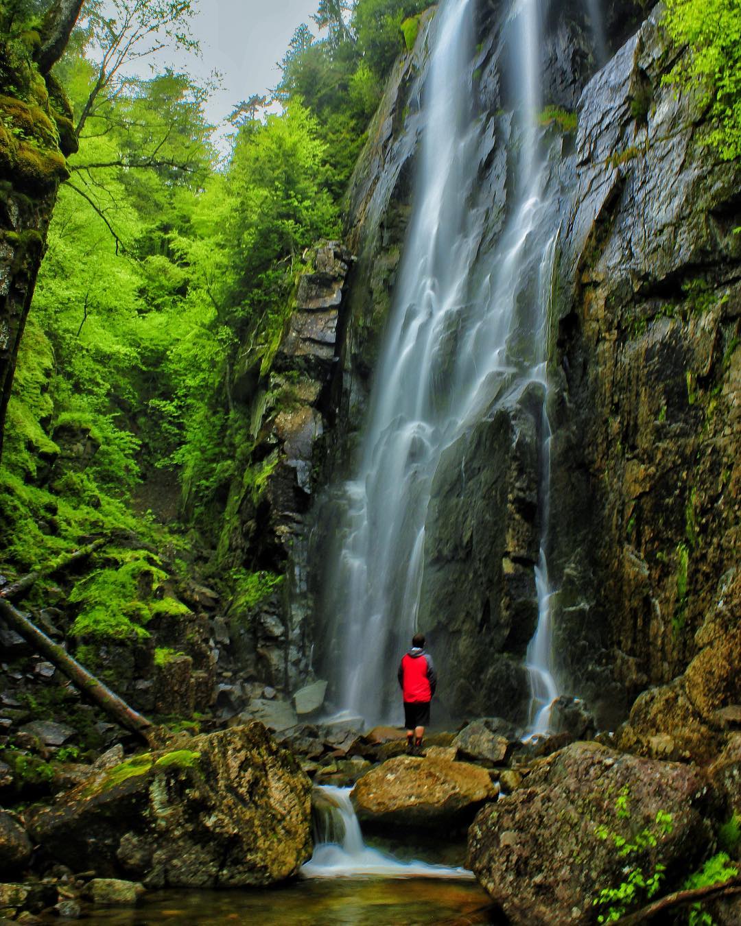A man facing backwards standing on a rock in front of Rainbow Falls.