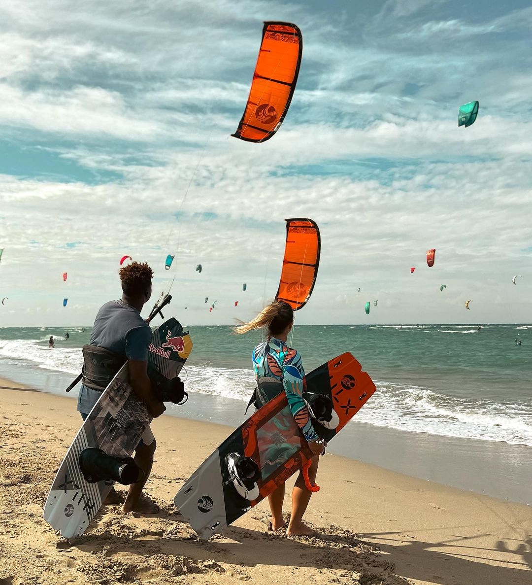 A man and woman holding kite surfing boards on the shore with people kitesurfing on water in the background at Kite Beach.
