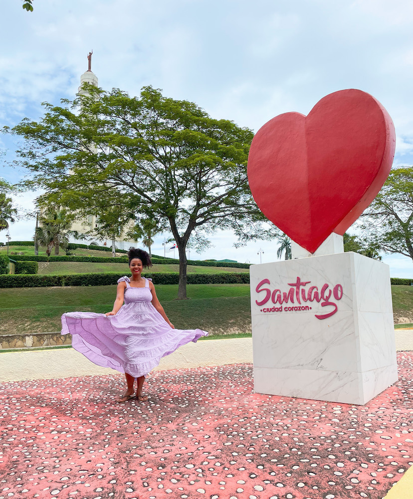 A woman in lilac dress standing beside a heart statue in Santiago, Dominican Republic.