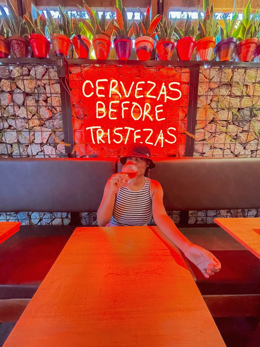 A woman sitting at a table drinking beer in front of a sign that says "cervezas before tristezas".