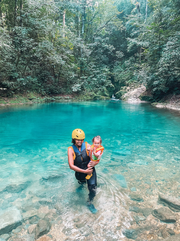 A woman carrying a baby at a natural pool surrounded by trees.