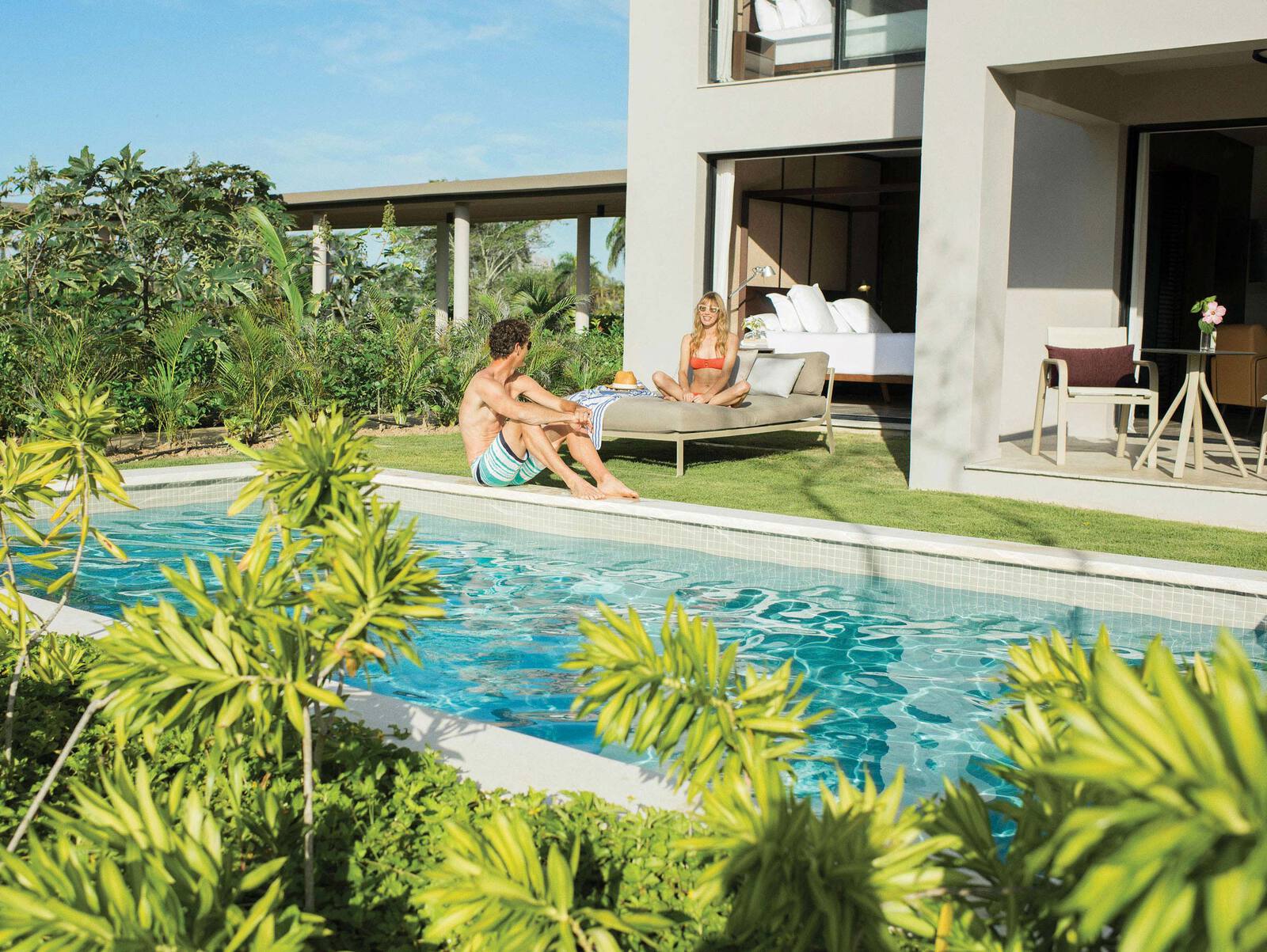 A woman sitting on an outdoor bed and a man sitting on the edge of a swimming pool at a resort in the Dominican Republic.
