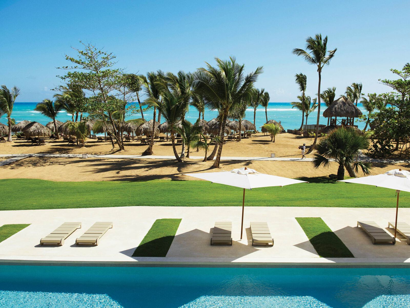 Outdoor swimming pool area by the sea with palm trees and huts at a resort in Uvero Alto, Punta Cana, Dominican Republic.