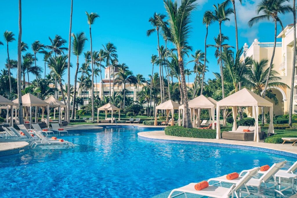 An outdoor swimming pool area with chairs and beds and palm trees at a resort in the Dominican Republic.