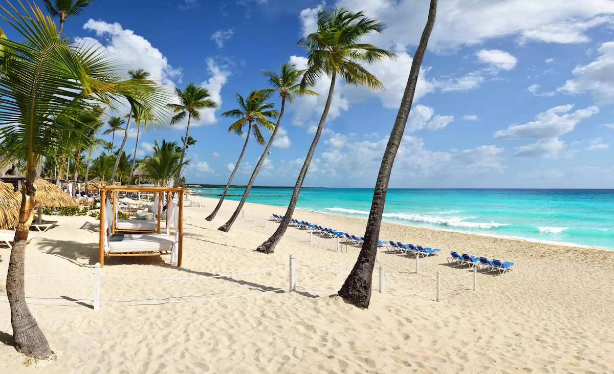A beach with blue waters, white sand, palm trees, chairs, and beds in La Romana, Dominican Republic.