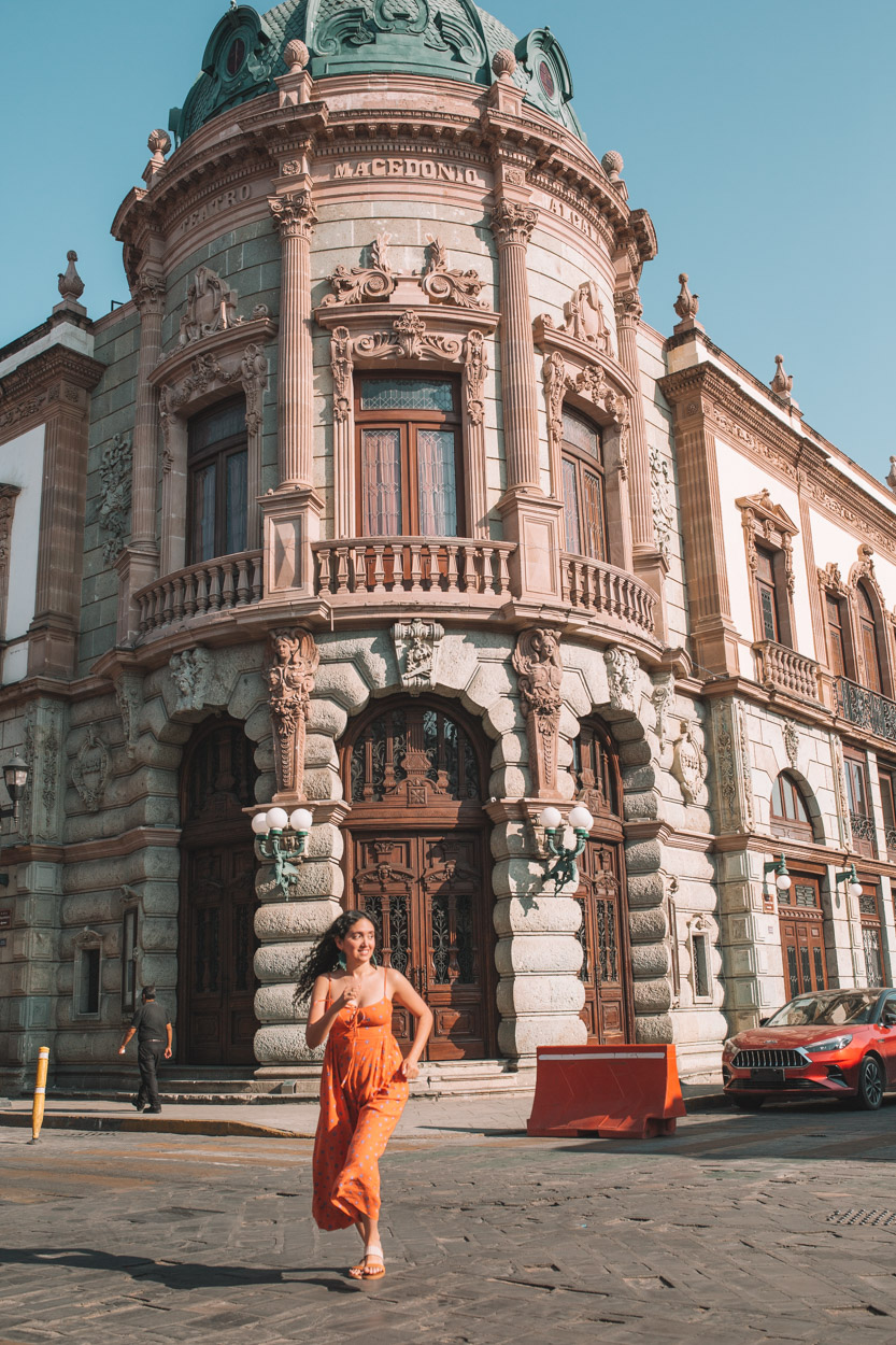 A woman in orange dress crossing a street in Oaxaca with an old building in the background.