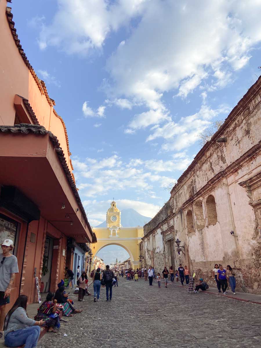 People walking and sitting on a sidewalk on Calle del Arco in Antigua with a volcano in the background