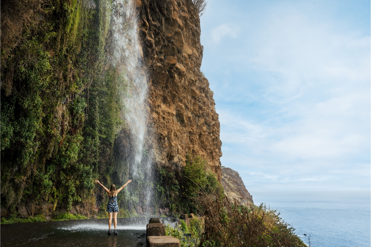 A woman standing on the road, with her arms spread out, and looking at the Cascata dos Anjos waterfall in Madeira
