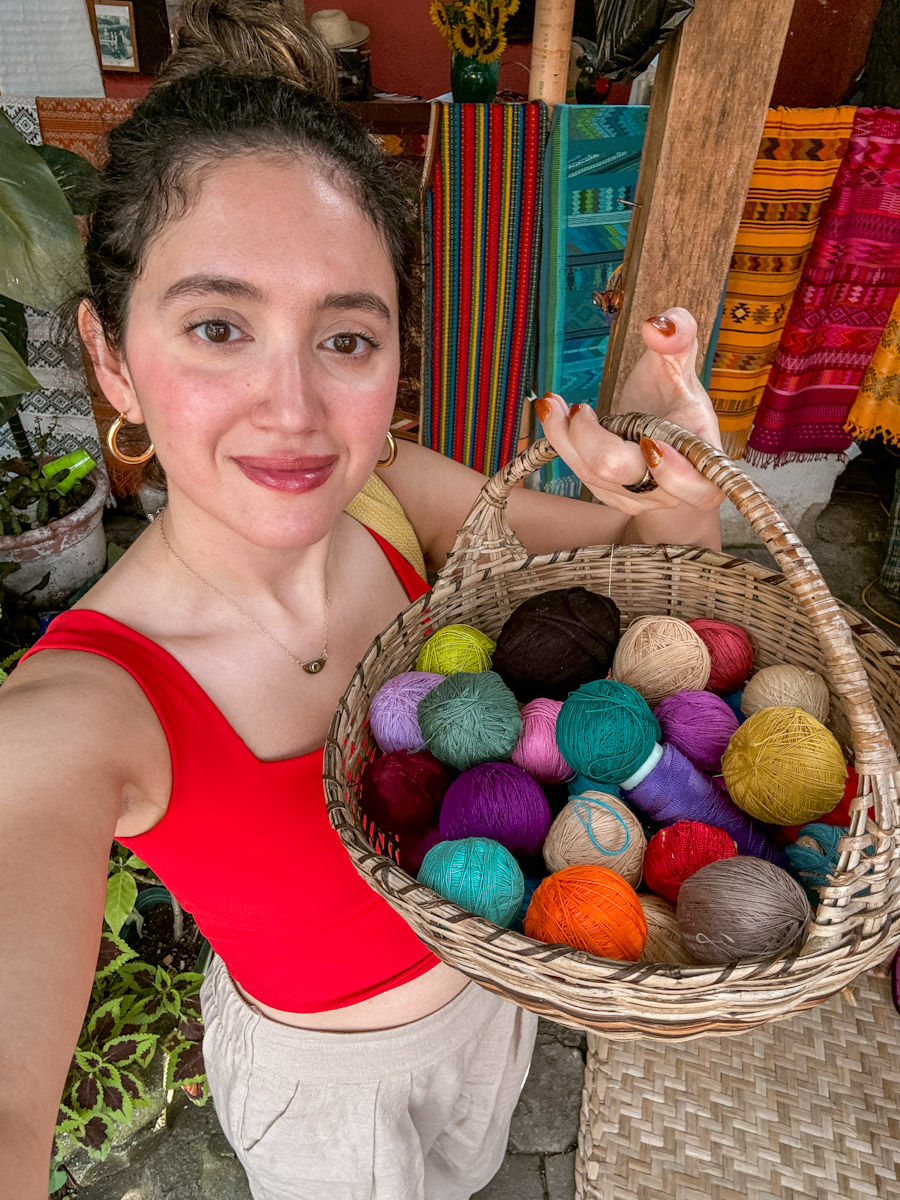 A woman holding a basket of yarns with textiles behind her.