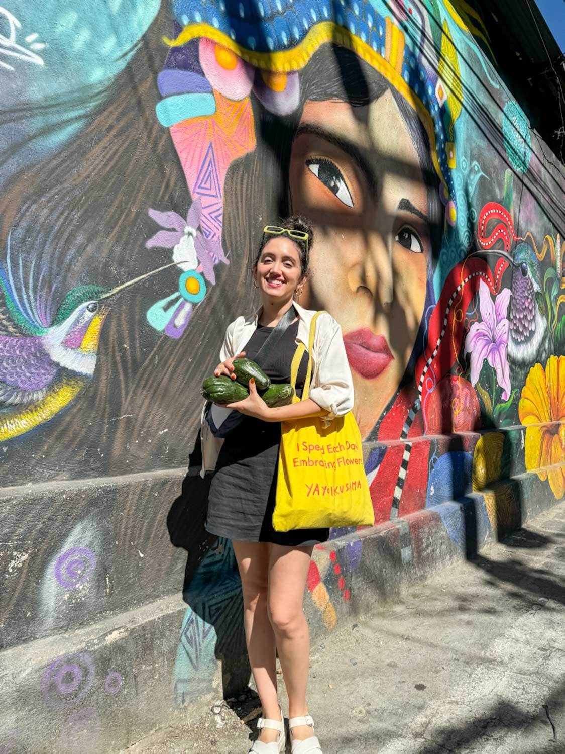 A woman standing in front of a colorful mural holding a yellow tote bag and avocadoes