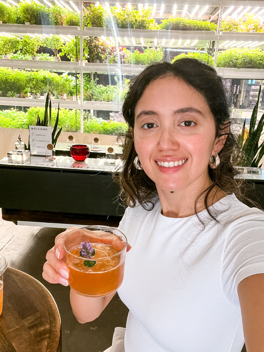 A woman holding a glass of cocktail with plants in the background