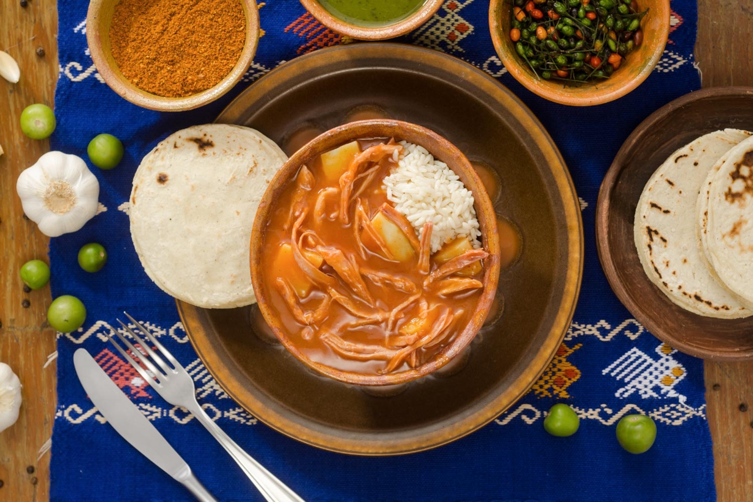 A bowl of hilachas on a table surrounded by utensils, tortillas, and other spices