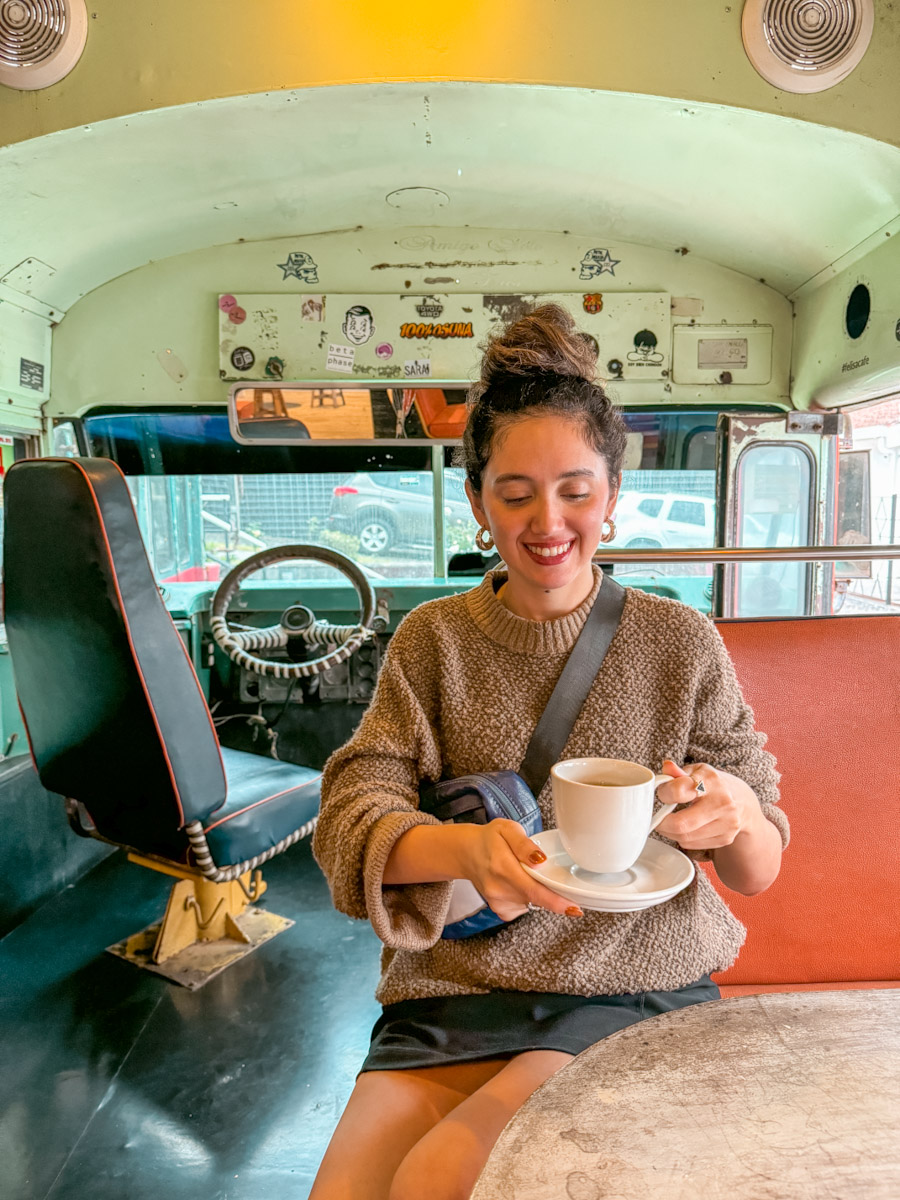 A woman holding a cup of coffee and a saucer inside a coffee shop with bus interior.
