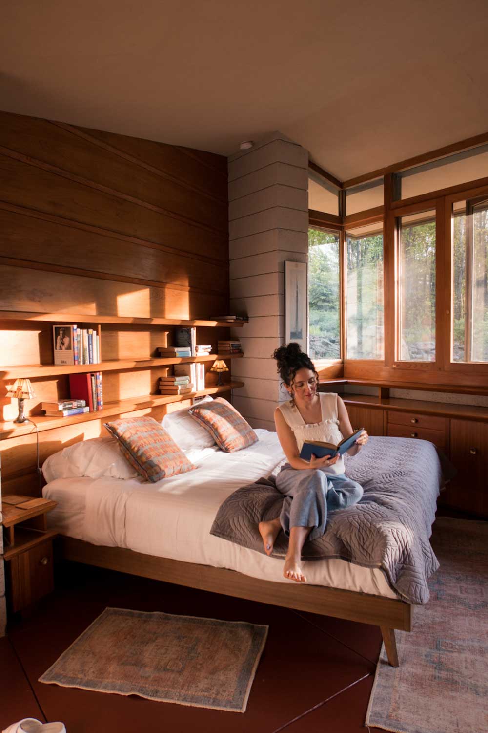 A woman sitting on a bed reading a book inside a wooden bedroom.