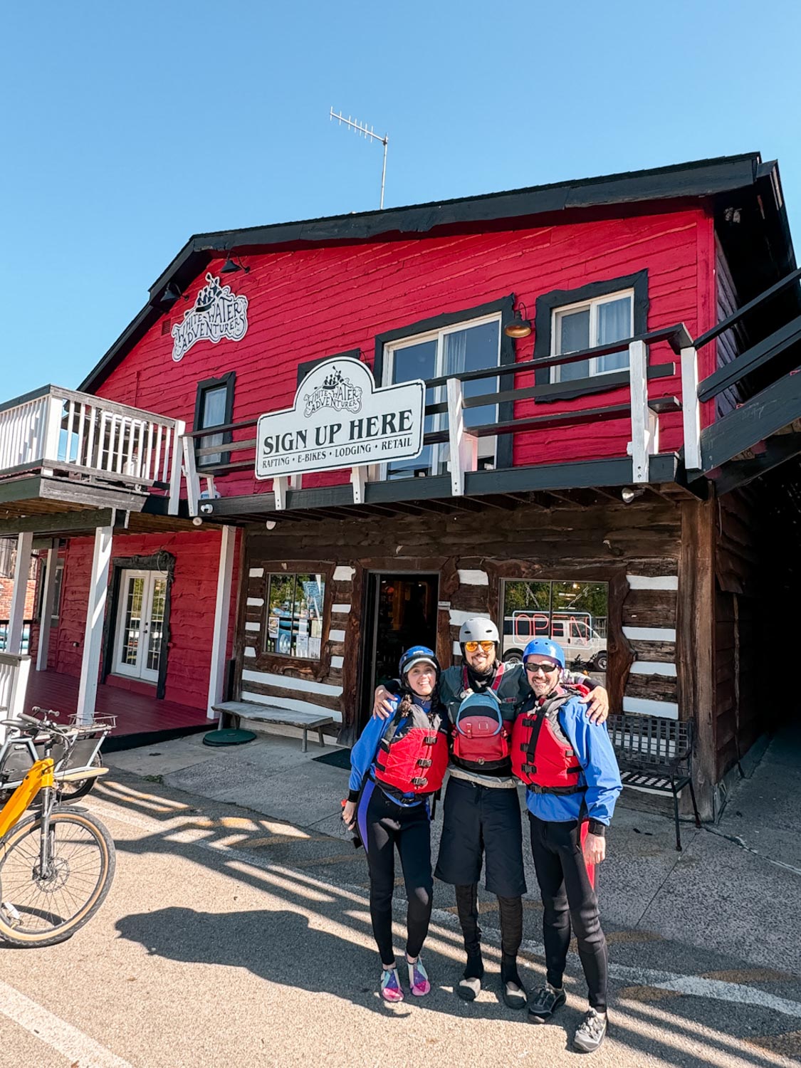 A woman and two men standing in front of a red wooden building wearing whitewater rafting gear.