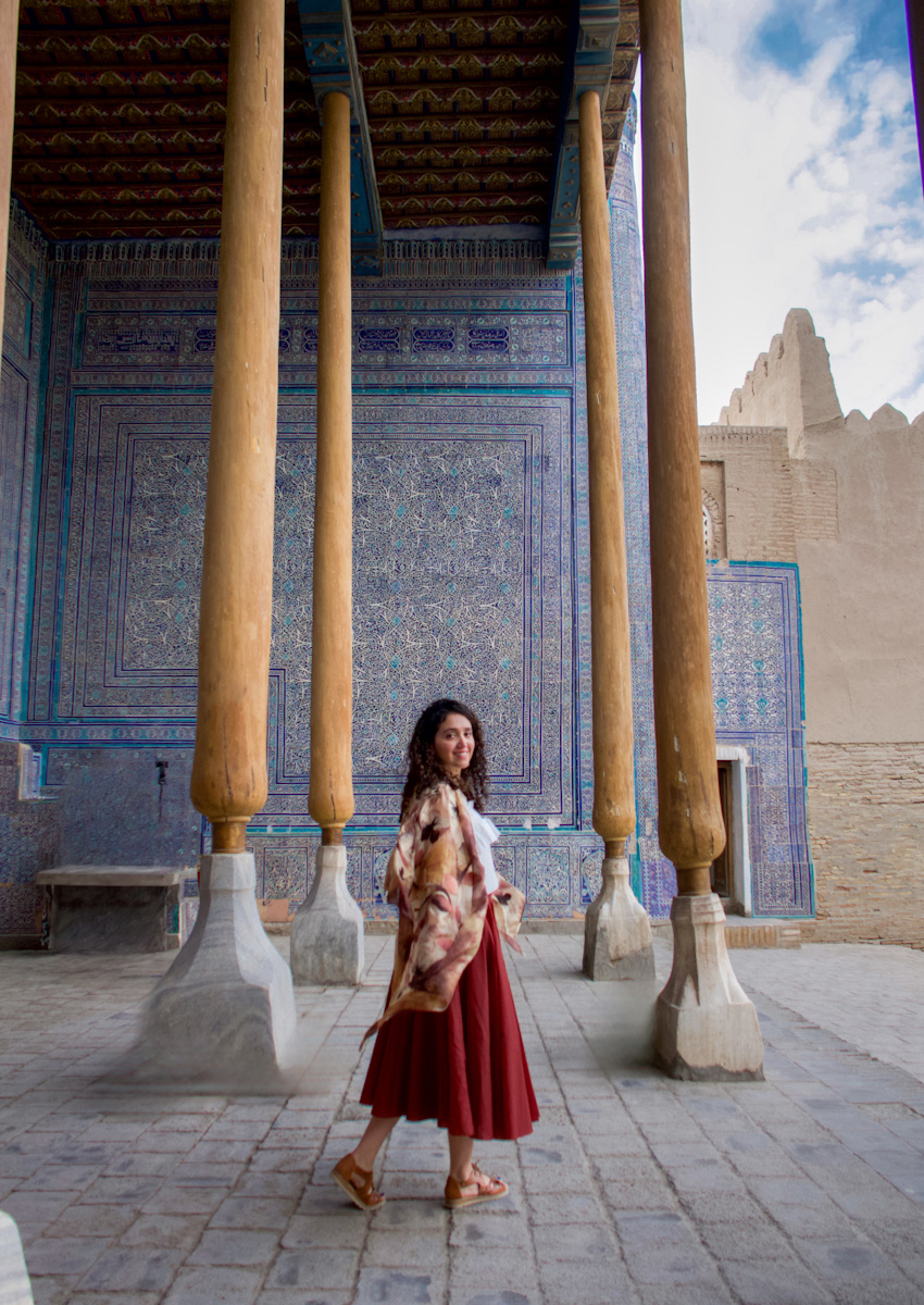 A woman standing in front of a wall with blue tile work and wooden pillars in Khiva.
