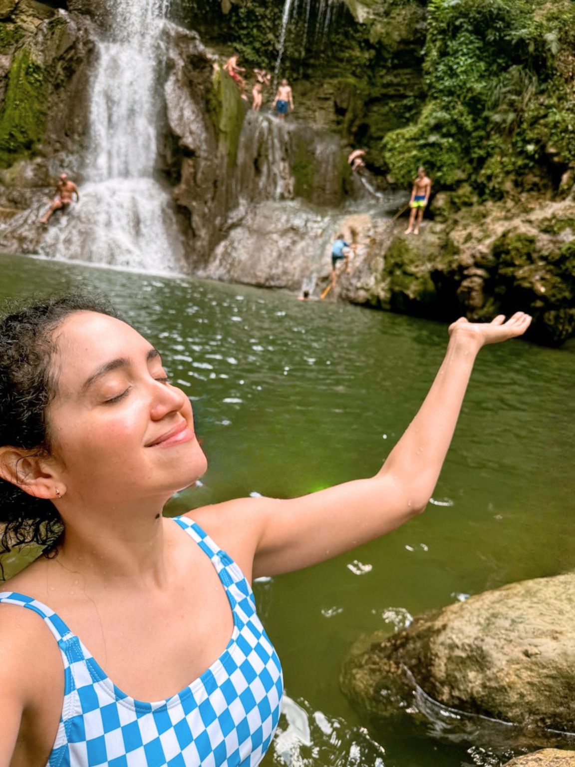 A woman in a swimsuit taking a selfie with people and a waterfall in the background.