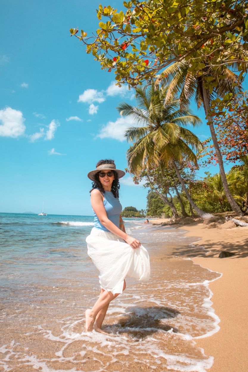 A woman wearing a hat, blue top, and long white skirt dipping her toes on the beach in Puerto Rico.