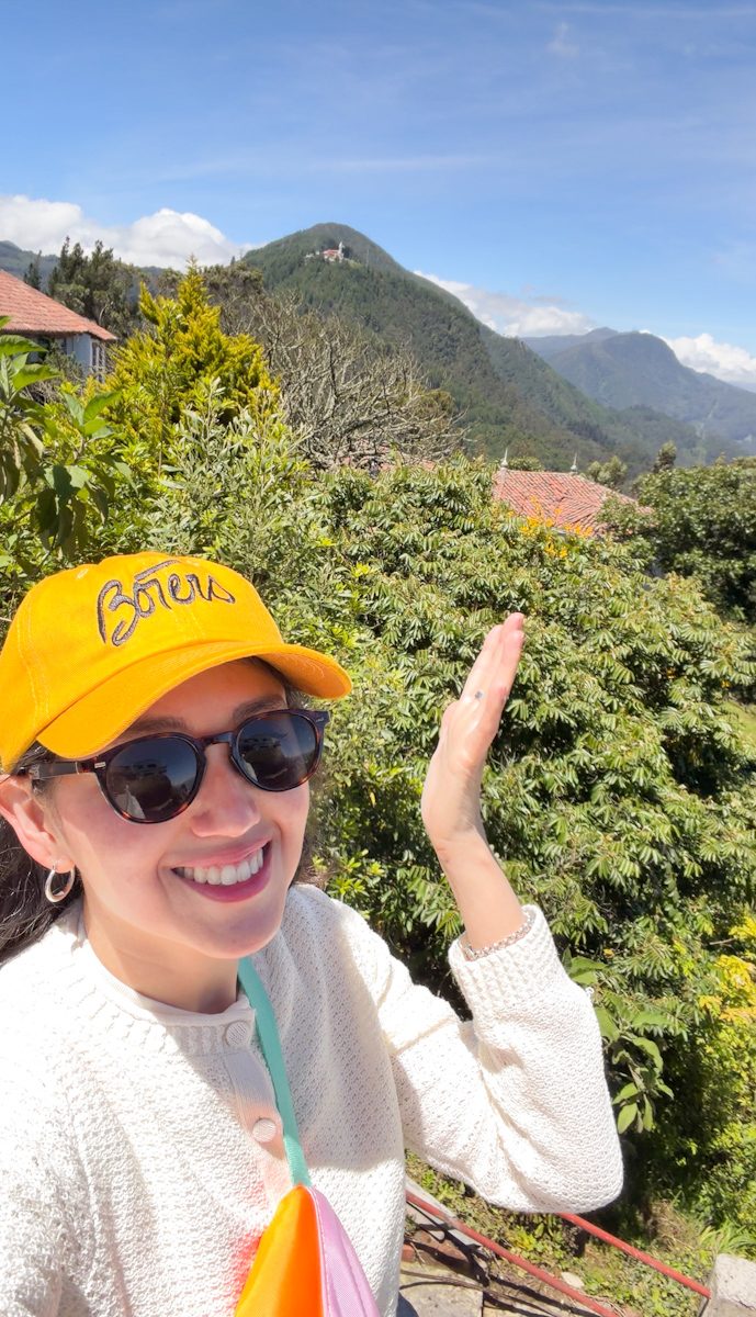 A woman in white top, black sunglasses, and yellow hat taking a picture in front of the Andes mountains in Bogota.