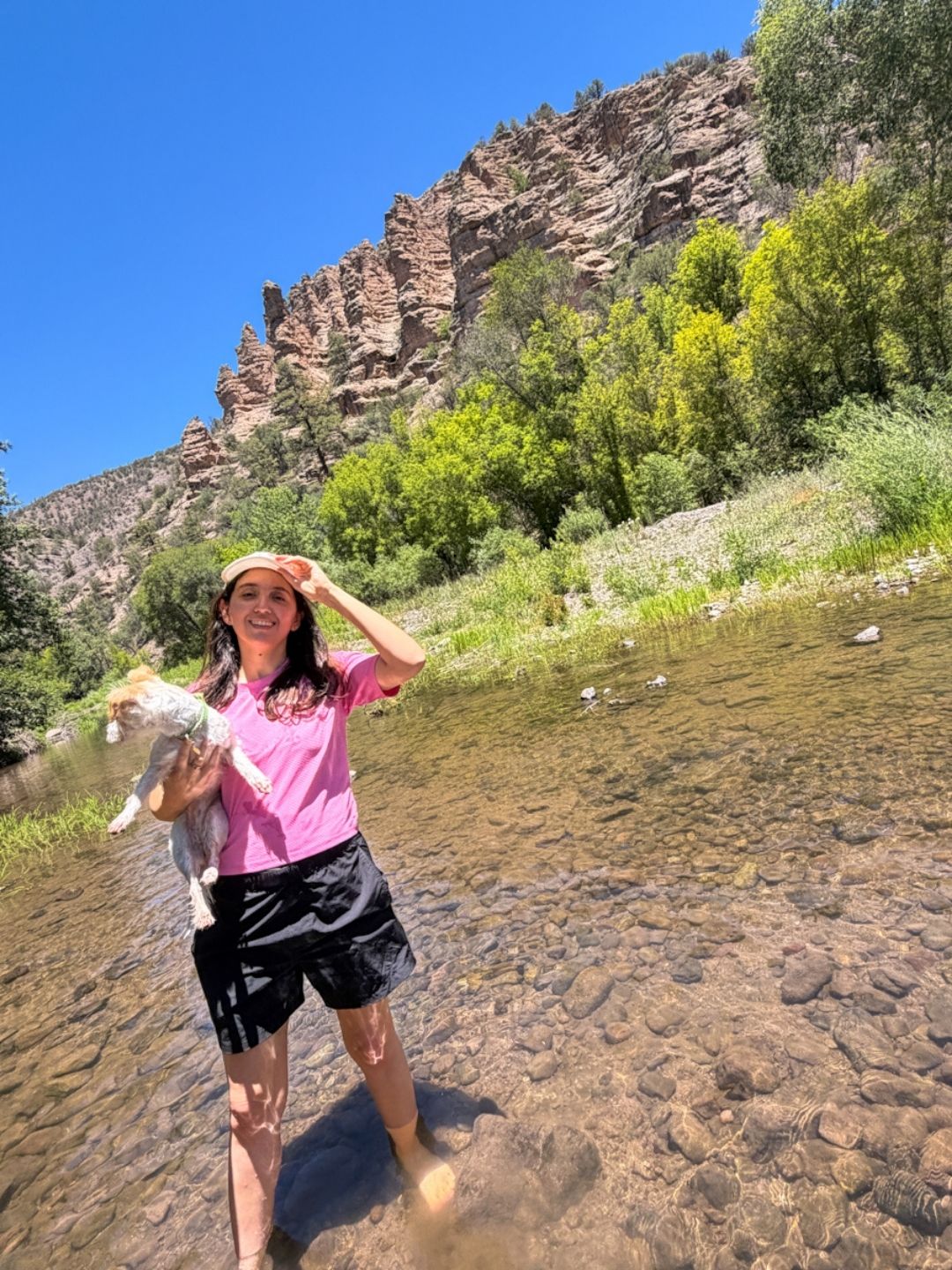 A woman holding a dog and standing on a hot springs with giant rock cliffs and trees in the background.