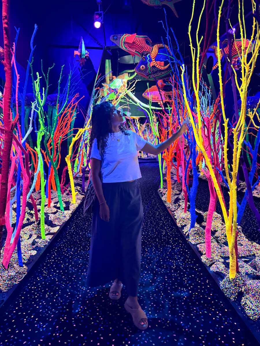 A woman standing inside a room with neon light colored underwater themed exhibit.