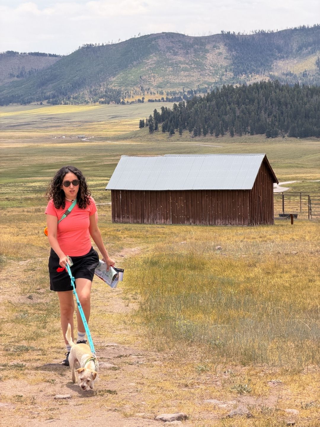 A woman holding a dog by its leash walking at a clearing with trees and a wooden building in the background in New Mexico.