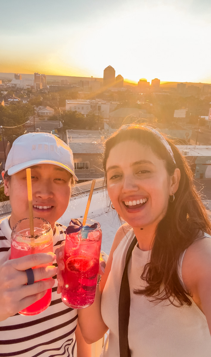 Two women holding glasses of drinks on a rooftop at sunset in Albuquerque, New Mexico.