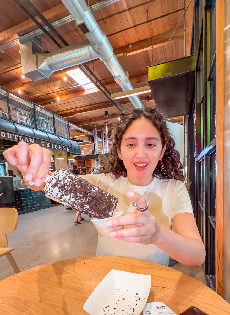A woman holding a stick of ice cream and dipping it in a cup.